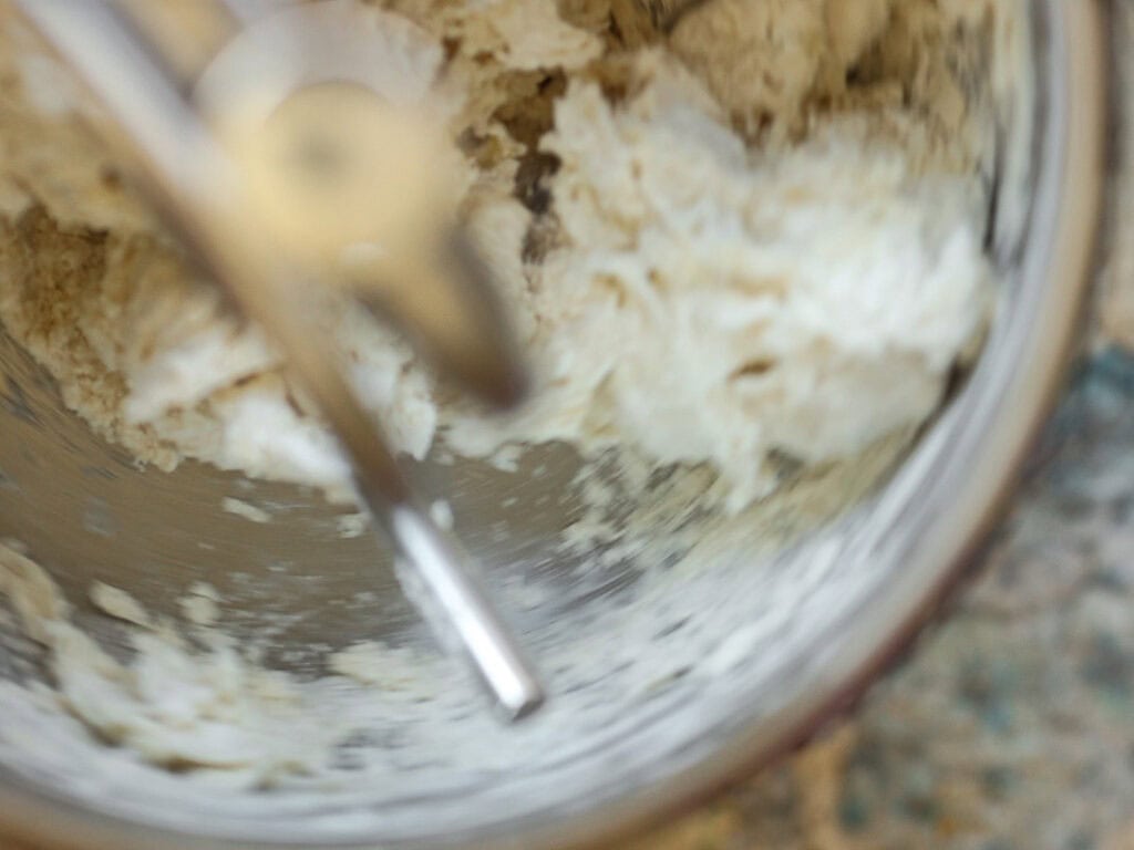 Close-up of dough in a metal mixing bowl, partially mixed by a silver dough hook. The dough is light in color and appears to be in the process of being kneaded. The background shows a speckled countertop.